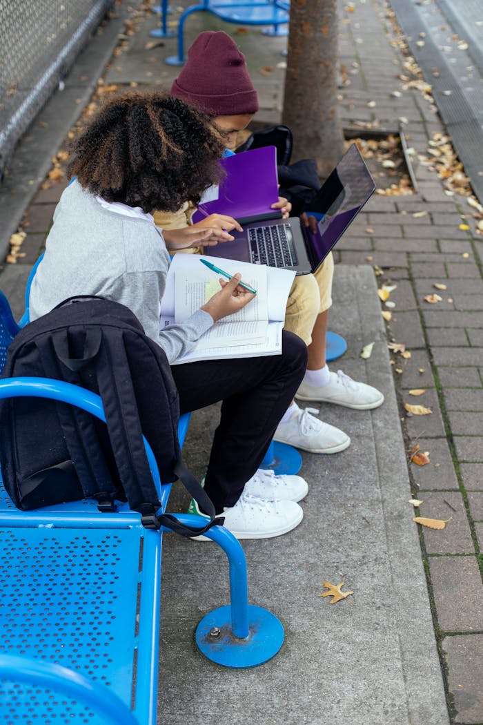 Anonymous multiethnic schoolchildren with laptop and copybooks studying on bench